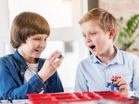 Surprised boy is looking at detail, his friend showing to him. They sitting near table in workshop